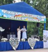 Scena plenerowa pod niebieskim namiotem; dwie osoby czytają z kartki, nad nimi baner „Piknik Integracyjny Razem Raźniej”, przed sceną dekoracje i kolaże zdjęć.