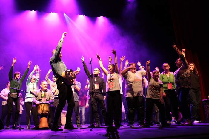 Pokaż zdjęcie: Zdjęcie sceny robione z dołu ku górze, na scenie tańczą młodzi ludzie, scena podświetlona jest na różowo, część młodzieży ma takie same szare koszulki z napisem akademia artystyczna, trzymają ręce uniesione do góry. Jedna z osób gra na gitarze, a jedna na bębnach.