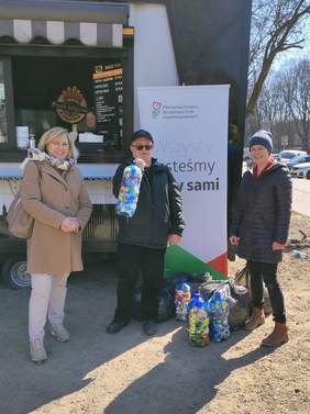 Pokaż zdjęcie: Przekazanie nakrętek zebranych przez pracowników Małopolskiego Oddziału PFRON. Trzy osoby stoją przed food truc`kiem, w rękach trzymają pojemniki, w których są kolorowe, plastikowe nakrętki.