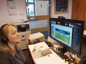 Pokaż zdjęcie: Kobieta w blond długich włosach z słuchawkami na uszach siedzi za biurkiem i wpatruje się w duży monitor. Na biurku leży rozłożony segregator z notatkami.