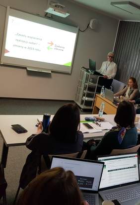 Pokaż zdjęcie: Grupa kilku osób siedzi za ustawionymi w dwóch rzędach stolikami. Część z tych osób posiada laptop ustawiony na stoliku, natomiast pozostała grupa ma przed sobą notatki w formie papierowej. Z przodu sali widoczne są dwie kobiety prowadzące szkolenie oraz ekran, na którym wyświetlana jest prezentacja.