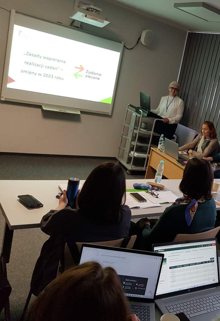 Pokaż zdjęcie: Grupa kilku osób siedzi za ustawionymi w dwóch rzędach stolikami. Część z tych osób posiada laptop ustawiony na stoliku, natomiast pozostała grupa ma przed sobą notatki w formie papierowej. Z przodu sali widoczne są dwie kobiety prowadzące szkolenie oraz ekran, na którym wyświetlana jest prezentacja.