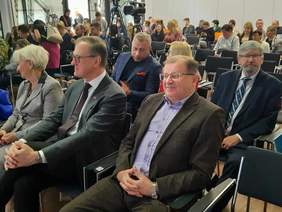 Pokaż zdjęcie: W rzędach siedzą na krzesłach ludzie. Są to uczestnicy konferencji. W tle widać kamerę, która filmuje wydarzenie. 