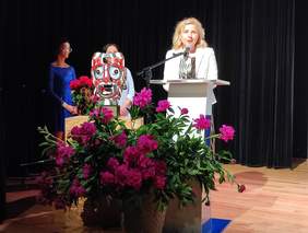 Pokaż zdjęcie: Na scenie przy mównicy stoi kobieta blondynka ubrana w czarno białą sukienkę oraz jasny żakiet. Przed mównicą stoi wazon z piwoniami