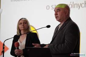 Pokaż zdjęcie: Na scenie przy mównicy stoi kobieta w granatowym garniturze oraz starszy mężczyzna, który mówi do mikrofonu.