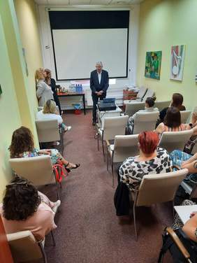 Pokaż zdjęcie: W sali siedzą na krzesłach ludzie. Ich sylwetki widać od tyłu. Przed nimi stoi pan w garniturze, który przemawia. Obok mężczyzny stoją dwie kobiety. 