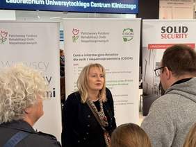 Pokaż zdjęcie: Stoisko informacyjne PFRON, CIDON w galerii, przy którym kobieta w czarnej marynarce rozmawia z grupą osób.