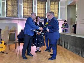 Pokaż zdjęcie: Na parkiecie stoi trzech mężczyzn w garniturach, pośrodku stoi kobieta w ciemnej sukni. Dwaj z nich podają sobie ręce jeden z przekazuje drugiemu nagrodę oprawioną w ramę drugi trzyma statuetkę w ręku.