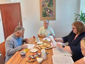 Pokaż zdjęcie: W jasnym pomieszczeniu, przy stole z poczęstunkiem siedzą 4 osoby. Dwóch mężczyzn pochyla się nad dokumentami do podpisu. Kobieta siedząca obok układa dokumenty.