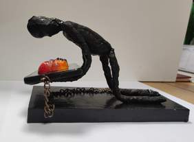 Pokaż zdjęcie: Zdjęcie rzeźby w kolorze czarnym przedstawiająca człowieka klęczącego nad telefonem i przykutego do niego łańcuchami. Na telefonie są trzy twarze z logo facebook, instagram i twitter