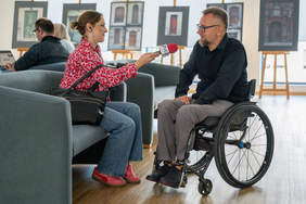 Kobieta, reporterka Radia PiK, siedząc na kanapie z mikrofonem w dłoni, przeprowadza wywiad z mężczyzną siedzącym na wózku, uczestnikiem konferencji i panelu dyskusyjnego  - Krzysztofem Kranzem