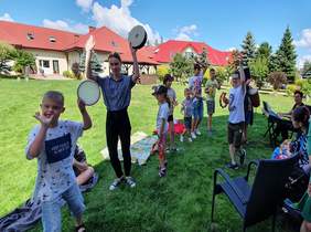 Pokaż zdjęcie: grupa dzieci bawi się na powietrzy w rękach trzymają instrumenty w oddali siedzi Pani i gra na instrumencie klawiszowym, wokoło dzieci śpiewają i tańczą 