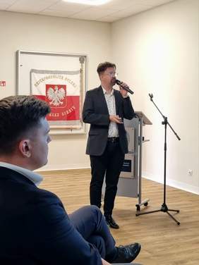 ​Opis alternatywny: Mężczyzna w ciemnej marynarce, jasnej koszuli we wzory i okularach stoi na środku sali i przemawia do mikrofonu, trzymając w drugiej ręce pilot do przerzucania slajdów. W tle, po lewej stronie, wisi oprawiona w ramę flaga z godłem Polski (białym orłem na czerwonym tle) oraz napisem „RZECZPOSPOLITA POLSKA”. Po prawej stronie widać pusty statyw mikrofonowy oraz mównicę. Na pierwszym planie, z boku i tyłu, widoczny jest siedzący z boku profil innego mężczyzny w ciemnym garniturze, który słucha wystąpienia. Podłoga w sali jest jasna, wykonana z paneli imitujących drewno.