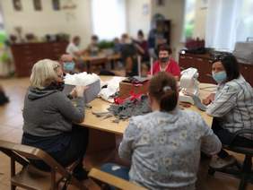 Pokaż zdjęcie: Panie siedzące przy stole wycinają z filcu elementy zabawek świątecznych. Oprócz ścinek i materiałów do wypychania stoi maszyna do szycia.