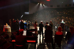 Grupa osób w odświętnych, wieczorowych strojach, na scenie, przygaszone światło, zdjęcie od tyłu