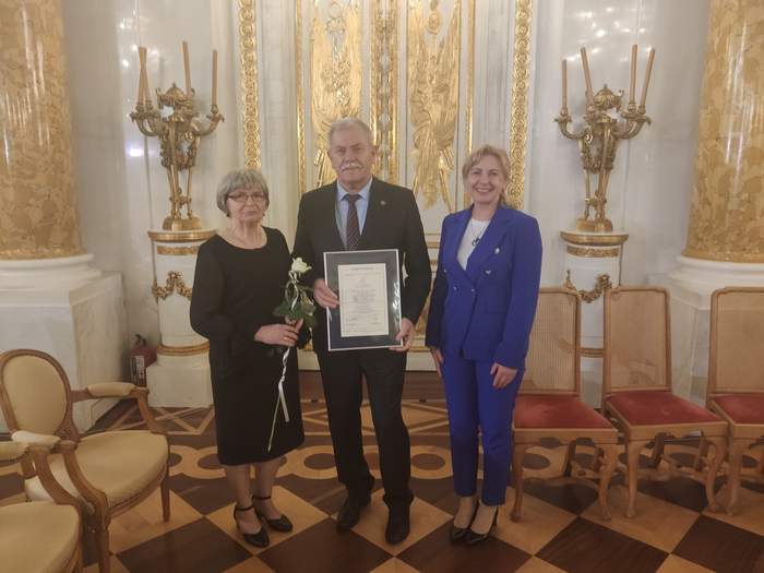 Pokaż zdjęcie: W sali na Zamku Królewskim w Warszawie pozują trzy osoby, dwie kobiety i mężczyzna. Mężczyzna stojący w środku trzyma dyplom