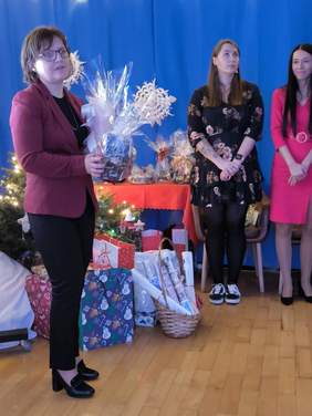 Pokaż zdjęcie:  Kobieta w bordowej marynarce stoi, trzymając w rękach zapakowany w celofan prezent. W tle widoczna jest choinka oraz inne osoby uczestniczące w wydarzeniu.