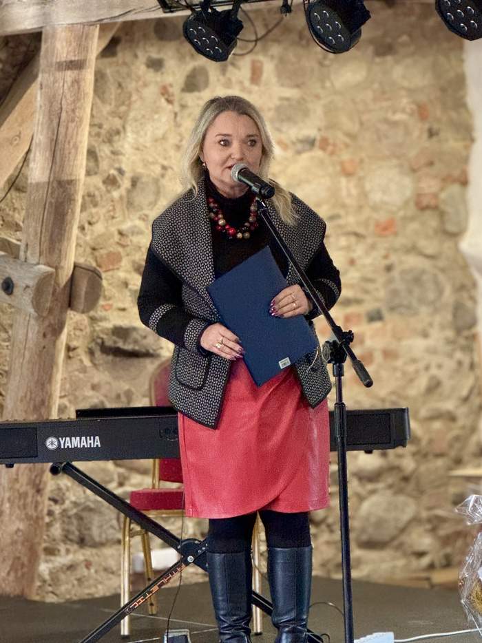 Pokaż zdjęcie: Kobieta ubrana w czarny sweter i czerwoną spódnicę w trakcie przemówienia do mikrofonu oraz trzyma w ręku dyplom, a za nią znajduje się klawiatura elektroniczna