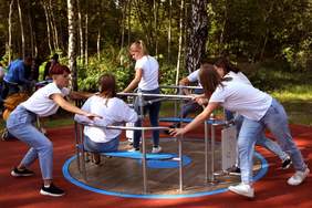 Pokaż zdjęcie: Na karuzeli kręcą się dzieci. Wszystkie mają białe koszulki. W tle widać drzewa. 