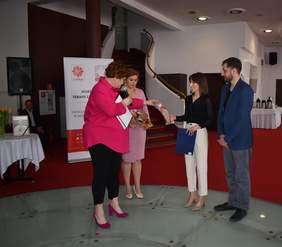 Pokaż zdjęcie: Cztery osoby uczestniczą w ceremonii wręczenia upominków. Kobieta w różowej marynarce przekazuje paczkę z prezentami kobiecie w czarnej bluzce i białych spodniach. Obok stoją druga kobieta w różowej sukience oraz mężczyzna w niebieskiej marynarce. W tle baner Caritas Diecezji Drohiczyńskiej.