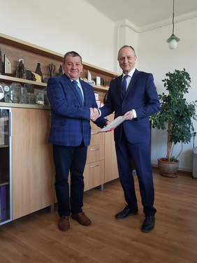 Dwaj mężczyźni w garniturach stoją, podają sobie dłonie, przekazują dokumenty. Uśmiechnięci, skierowanie w stronę robiącego zdjęcie.