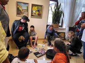 Pokaż zdjęcie: Artyści malują nogami. Dookoła siedzą uczestnicy spotkania, którzy obserwują jak powstają Ich prace.