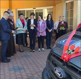 Pokaż zdjęcie: Grupa osób pozuje przy nowym, ośmioosobowym busie zaparkowanym przed budynkiem. Samochód zdobi czerwona wstążka na masce oraz czerwone tulipany. W tle jasna elewacja z dużymi oknami.