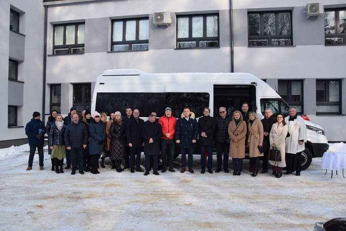 Pokaż zdjęcie: Przy białym autobusie stoi w rzędzie duża grupa osób, na dworze jest zimno, lezy śnieg, osoby sa ubrane płaszcze lub kurtki.