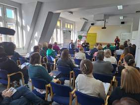 Uczestnicy konferencji, siedzą na krzesłach, widok od tyłu, z przodu widoczna jest prelegentka i banery