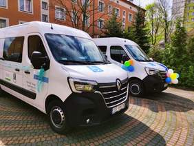 Pokaż zdjęcie: Obok siebie stoją dwa białe busy jeden jest obwiązany wstążką drugi jest ozdobiony różnokolorowymi balonami w tle fragmenty budynków