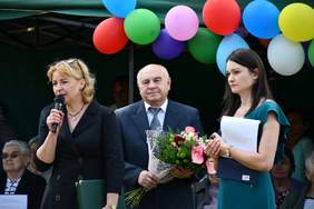 Pokaż zdjęcie: Trzy osoby, dwie kobiety i mężczyzna. Kobieta z lewej mówi do mikrofonu, mężczyzna stojący w środku trzyma kwiaty
