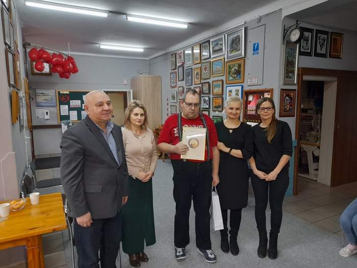 Pokaż zdjęcie: W pracowni artystycznej Warsztatu Terapii Zajęciowej pozuje do pamiątkowego zdjęcia 5 osób. Pośrodku stoi mężczyzna w czerwonej koszulce, w ręku trzyma dyplom. Po lewej stronie zdjęcia stoją pracownicy PFRON mężczyzna i kobieta, po prawej dwie kobiety pracujące w WTZ w Chełmie.