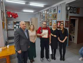 Pokaż zdjęcie: W pracowni artystycznej Warsztatu Terapii Zajęciowej pozuje do pamiątkowego zdjęcia 5 osób. Pośrodku stoi mężczyzna w czerwonej koszulce, w ręku trzyma dyplom. Po lewej stronie zdjęcia stoją pracownicy PFRON mężczyzna i kobieta, po prawej dwie kobiety pracujące w WTZ w Chełmie.