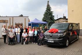 Pokaż zdjęcie: Grupa nieco ponad dwudziestu osób stoi przed niskim budynkiem, obok czarnego busa udekorowanego czerwoną kokardą. Wśród zgromadzonych osób, są trzy osoby, które poruszają się na wózku. W tle namiot, wysoka jodła oraz inne zabudowania.