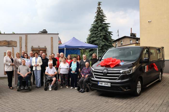 Pokaż zdjęcie: Grupa nieco ponad dwudziestu osób stoi przed niskim budynkiem, obok czarnego busa udekorowanego czerwoną kokardą. Wśród zgromadzonych osób, są trzy osoby, które poruszają się na wózku. W tle namiot, wysoka jodła oraz inne zabudowania.