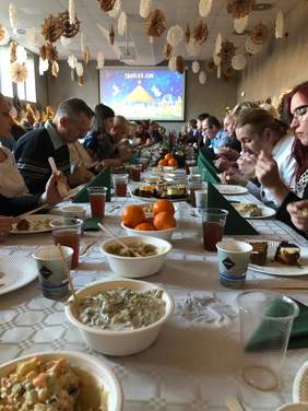 Pokaż zdjęcie: W sali konferencyjnej stoi długi stół. Wokół niego siedzą ludzie i jedzą świąteczne potrawy. Wszędzie jest dużo dekoracji bożonarodzeniowych. 