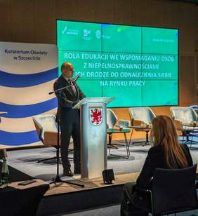 Pokaż zdjęcie: Zdjęcie przedstawia mężczyznę w ciemnym garniturze stojącego za mównicą na podwyższeniu w sali konferencyjnej. Na mównicy widnieje herb (czerwony gryf). Na dużym ekranie za nim wyświetlany jest tytuł prezentacji w języku polskim: "ROLA EDUKACJI WE WSPOMAGANIU OSÓB Z NIEPEŁNOSPRAWNOŚCIAMI ICH DRODZE DO ODNALEZIENIA SIEBIE NA RYNKU PRACY". W prawym górnym rogu ekranu widnieje data "SZCZECIN 21.10.2023" oraz logo "Pomorze Zachodnie" i inne. Po lewej stronie mównicy widać baner z napisem "Kuratorium Oświaty w Szczecinie" oraz fragment flagi Unii Europejskiej. Na pierwszym planie, tyłem do obiektywu, siedzi kobieta z długimi, jasnymi włosami.