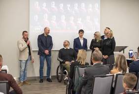 Pokaż zdjęcie: Zdjęcie przedstawia grupę osób na scenie w trakcie oficjalnego wydarzenia. W tle widoczny jest slajd prezentacyjny z fotografiami członków drużyny sportowej w czerwonych koszulkach. Na pierwszym planie znajdują się widzowie siedzący na krzesłach. Wśród osób znajdujących się na scenie jedna siedzi na wózku inwalidzkim a mężczyzna po prawej stronie mówi do mikrofonu reszta grupy się przysłuchuje. 