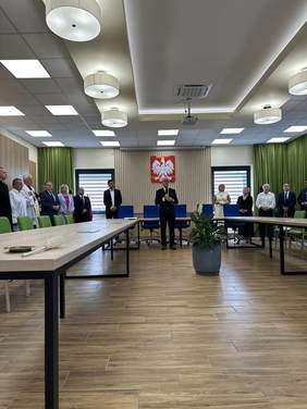 Pokaż zdjęcie: Sala konferencyjna ze stolikami, na ścianie wisi godło Polski, pod ścianą stoją zaproszeni gości.