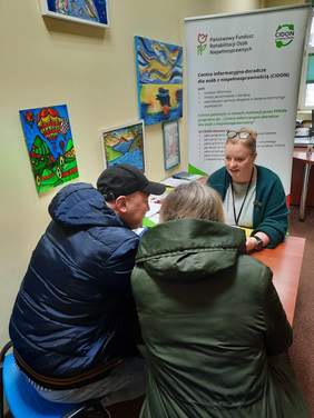 Pokaż zdjęcie: Przy biurku siedzi kobieta - ekspertka z Centrum Informacyjno-Doradczego dla Osób z Niepełnosprawnością. Naprzeciwko siedzi para - pani i pan. Eksperka odpowiada na pytania i uśmiecha się. Na ścianie w tle wiszą kolorowe obrazki. 