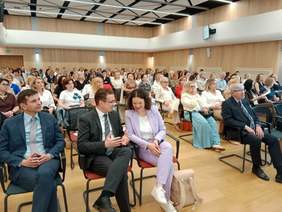 Pokaż zdjęcie: W auli uniwersyteckiej są rzędy krzeseł, na których siedzą ludzie. Wszyscy sluchają prelekcji. 