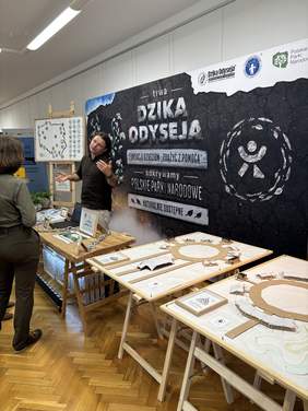 Pokaż zdjęcie: W sali znajdują się stoły z informatorami i modelami. W tle stoi duży czarny baner z napisem „DZIKA ODYSSEJA” i grafiką wilka. Przy banerze stoi kilka osób.
