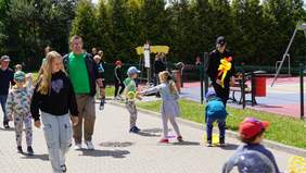 Pokaż zdjęcie: Na chodniku bawią się dzieci. W tle widać także plac zabaw. Niektóre dzieci trzymają zabawki, a inne balony.  