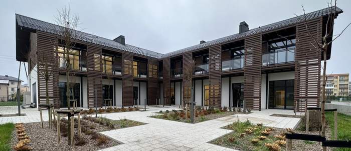 Pokaż zdjęcie: Symetryczny budynek w kształcie litery L. Na dole białe ściany u góry ściany wyłożone ciemnym drewnem. Szklane bariery balkonowe. Od dachu w dół symetrycznie schodzą ciemne drewniane pergole. 