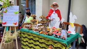 Pokaż zdjęcie: Za Stołem pełnym ciasta stoi kobieta przebrana w strój czerwonego kapturka. Obok sztaluga z napisem w Stawikowych lasach czerwony kapturek z pełnym koszyczkiem hasa.