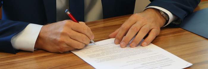 Pokaż zdjęcie: Mężczyzna ubrany w garnitur, białą koszulę i krawat podpisuje dokument. Używa długopisu. 