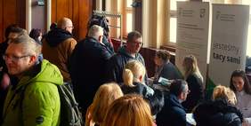 Pokaż zdjęcie: Dwie kobiety znajdują się przy stoisku Oddziału Wielkopolskiego PFRON, z czego jedna z nich siedzi na tle baneru CIDON, a druga na tle baneru PFRON. Przy stoisku obecna jest również duża grupa osób, które przyszły na Giełdę Pracy dla osób z niepełnosprawnością. Na stoliku ułożone zostały materiały informacyjne promujące programy i zadania PFRON. W pobliżu widoczne są także stoiska innych pracodawców.