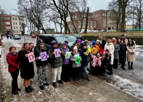 Pokaż zdjęcie: Grupa osób, składająca się z młodzieży i dorosłych, pozuje na zewnątrz. Większość osób jest ciepło ubrana w kurtki, czapki i szaliki. Centralnie przed ciemnym Fordem Transitem Custom trzymają kolorowe litery tworzące słowo "DZIĘKUJEMY". W tle, po prawej stronie, widać duży, zabytkowy budynek.