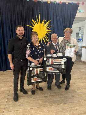 Pokaż zdjęcie: Cztery osoby uśmiechają się do aparatu. Dwie z nich – mężczyzna i kobieta trzymają zestawy plastyczne. Osoba znajdująca się po prawej stronie pokazuje dyplom. Za nimi ciemna kotara z żółtym słońcem na środku.