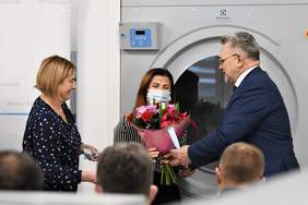 Pokaż zdjęcie: Na tle dużej pralki stoją dwie kobiety i mężczyzna, który wręcza jednej z kobiet bukiet kwiatów